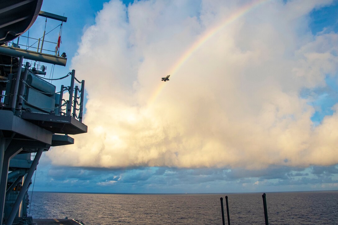 A fighter jet flies overhead by a rainbow against a backdrop of dramatic clouds over the ocean, with a military ship on the left.