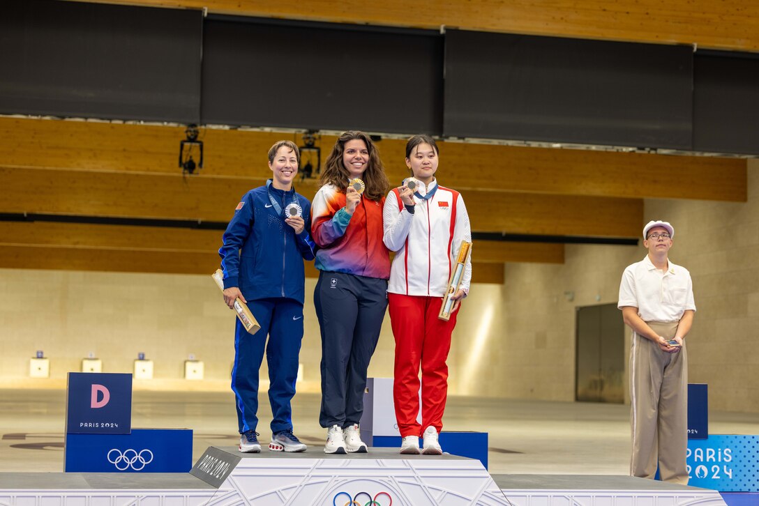 Three athletes in front of an indoor shooting range stand on a podium side by side holding bronze, silver and gold medals while an official stands on the right.