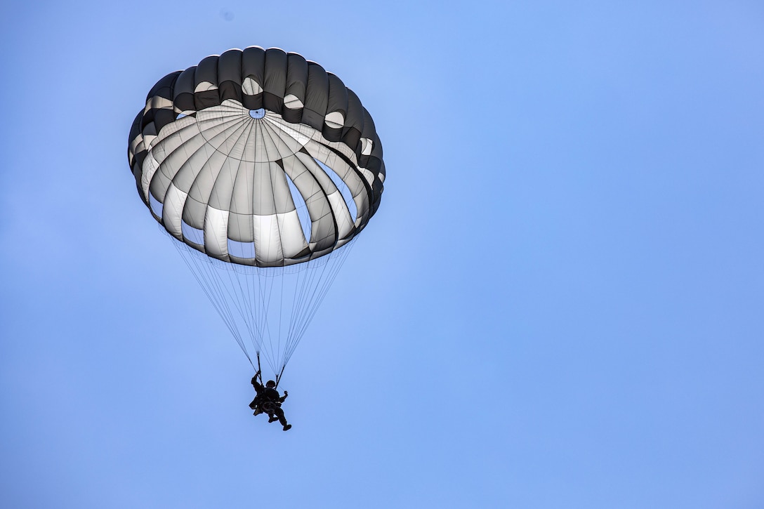 A person descends with a black and white parachute against a clear blue sky.