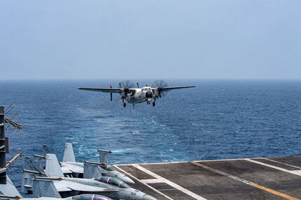 An aircraft approaches the landing deck of an aircraft carrier in open water during daylight.