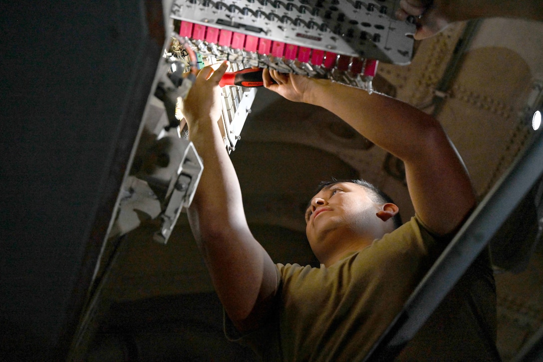 An airman uses a tool while working on an aircraft illuminated by an overhead light.