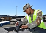 A Black soldier holds a small hatch open on the top of a metal container. he is outside.