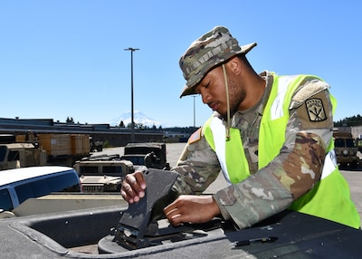 A Black soldier holds a small hatch open on the top of a metal container. he is outside.
