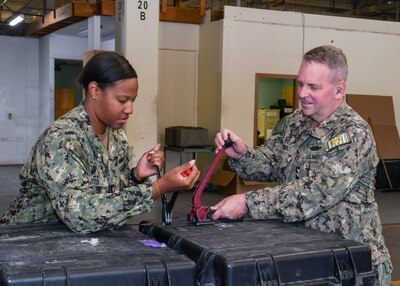 Two sailors, one is a Black woman the other a white male, are using equipment to band cargo containers. they are working in a warehouse.