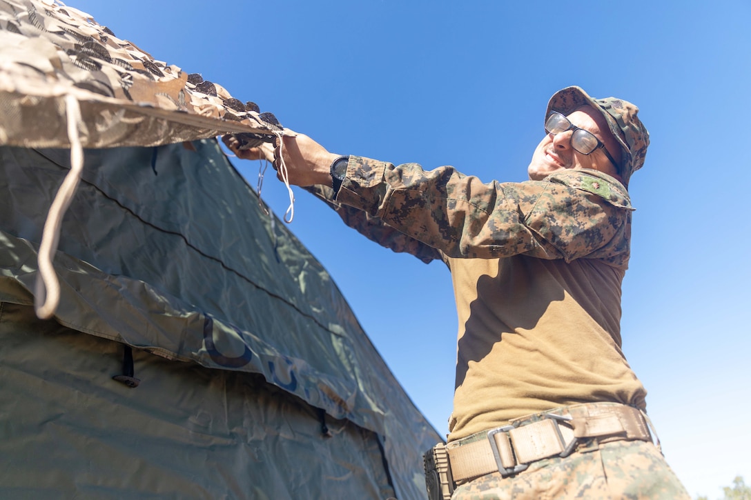 A sailor pulls a camo cover over a tent while standing in a field under a blue sky.