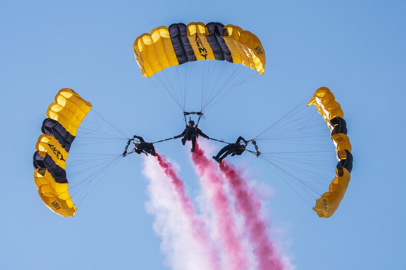 Three skydivers parachute while joined conjoined with streaks of pink smoke in their wake.
