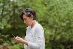 Woman wearing white sweater stands outside and looks at smartphone.