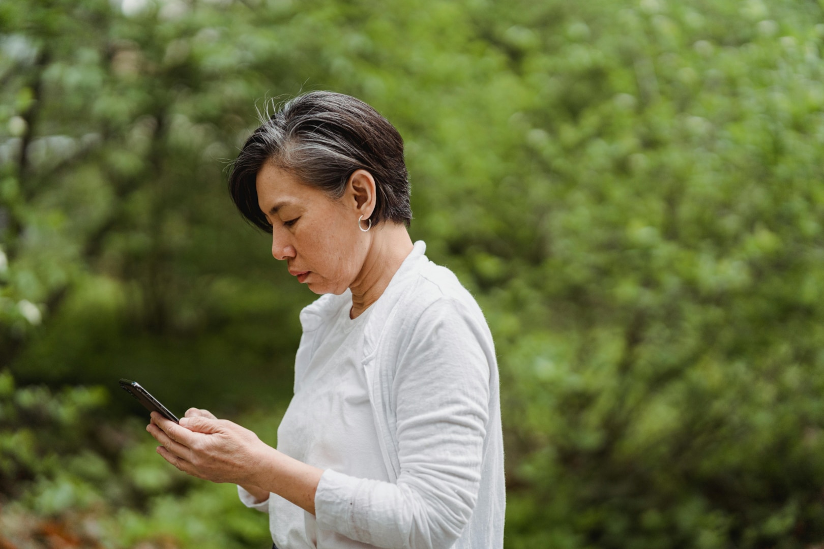 Woman wearing white sweater stands outside and looks at smartphone.