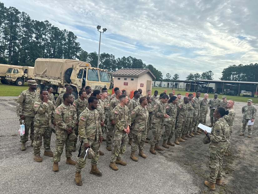 A group of soldiers receives orders from another soldier with vehicles parked in the background.