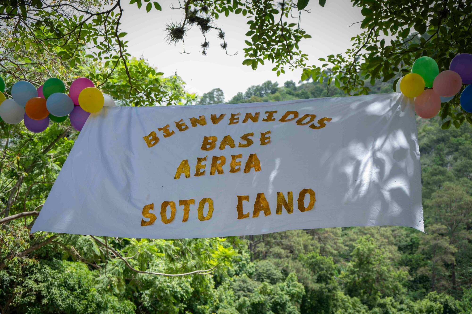 A sign that says "Bienvenidos Base Aerea Soto Cano" waves in the wind with balloons attached.