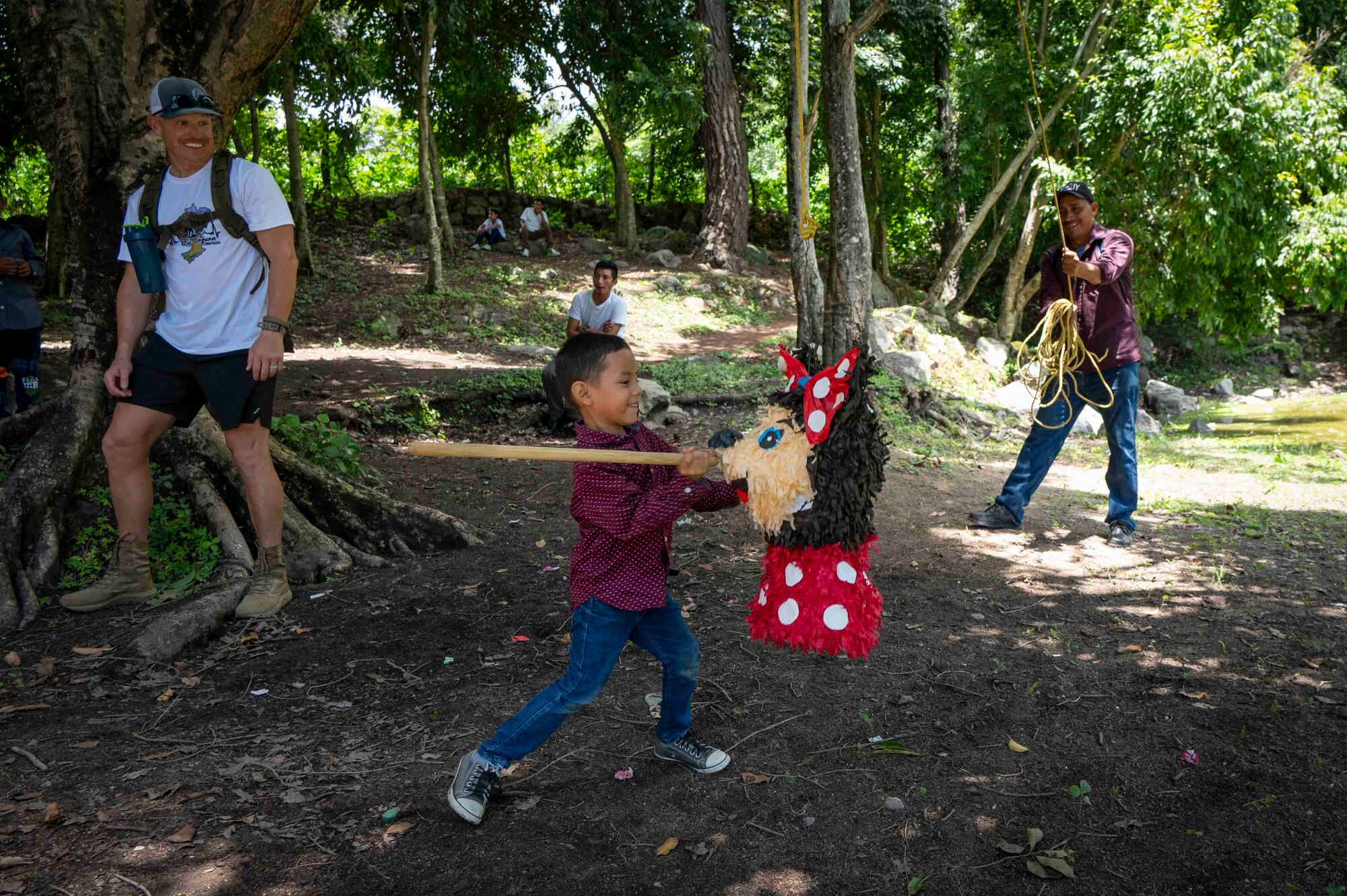A person smiles as he watches a child swing a stick at a piñata and another person smiles while holding the piñata rope.