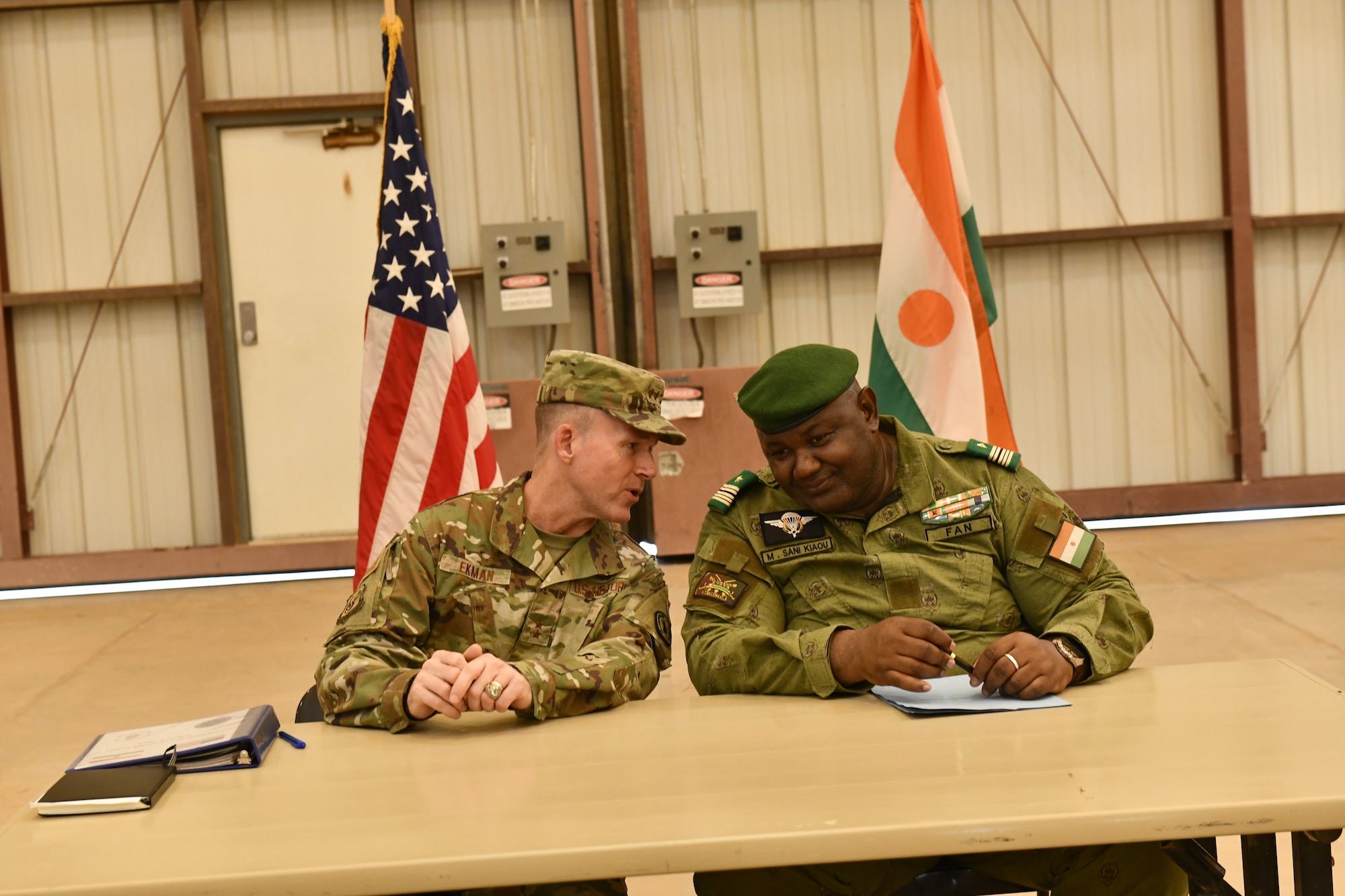 Two military members talk at a table in front of an American flag and a Nigerien flag