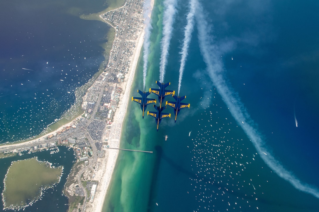 Four military jets fly above a narrow stretch of beach with boats dotting the water on both sides.