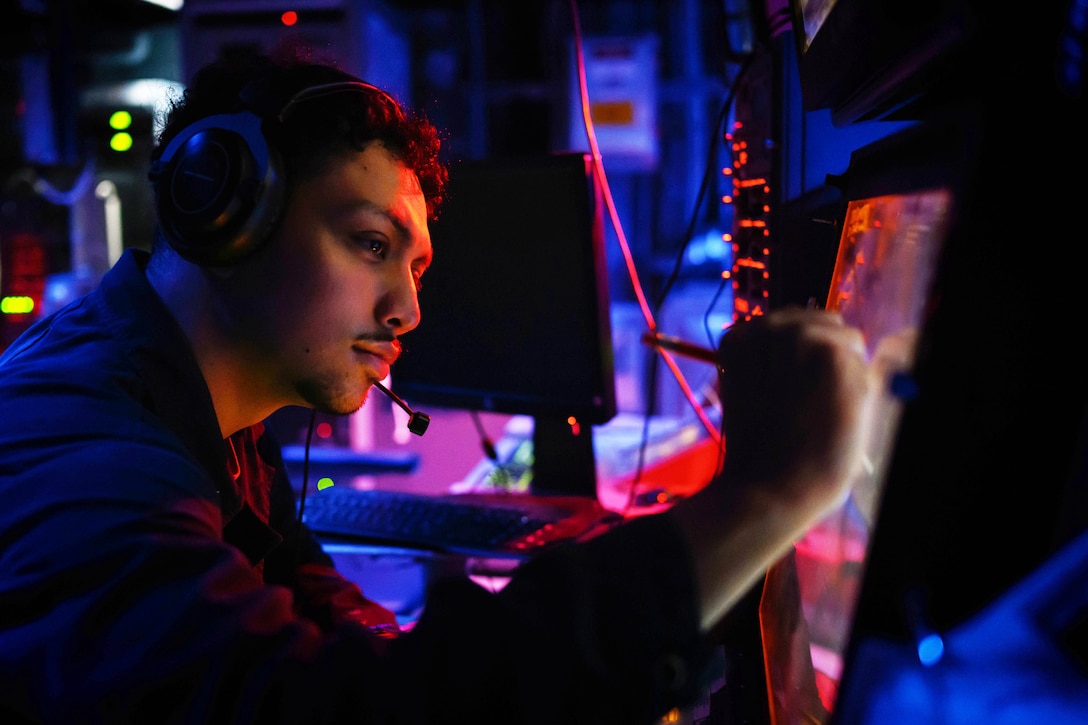 A sailor wearing a headset uses a stylus while focusing on a monitor in a dark room with red and blue lighting.