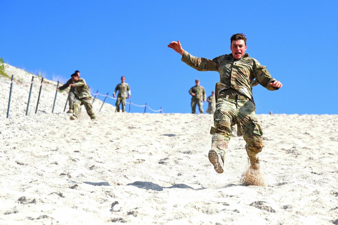 A soldier shouts while running down a sand dune. Other soldiers hold on to a rope barrier as they make their way down in the background.