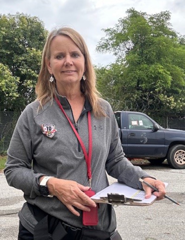 Photo of a woman in work clothes holding a clipboard looking at the camera, she is outside.