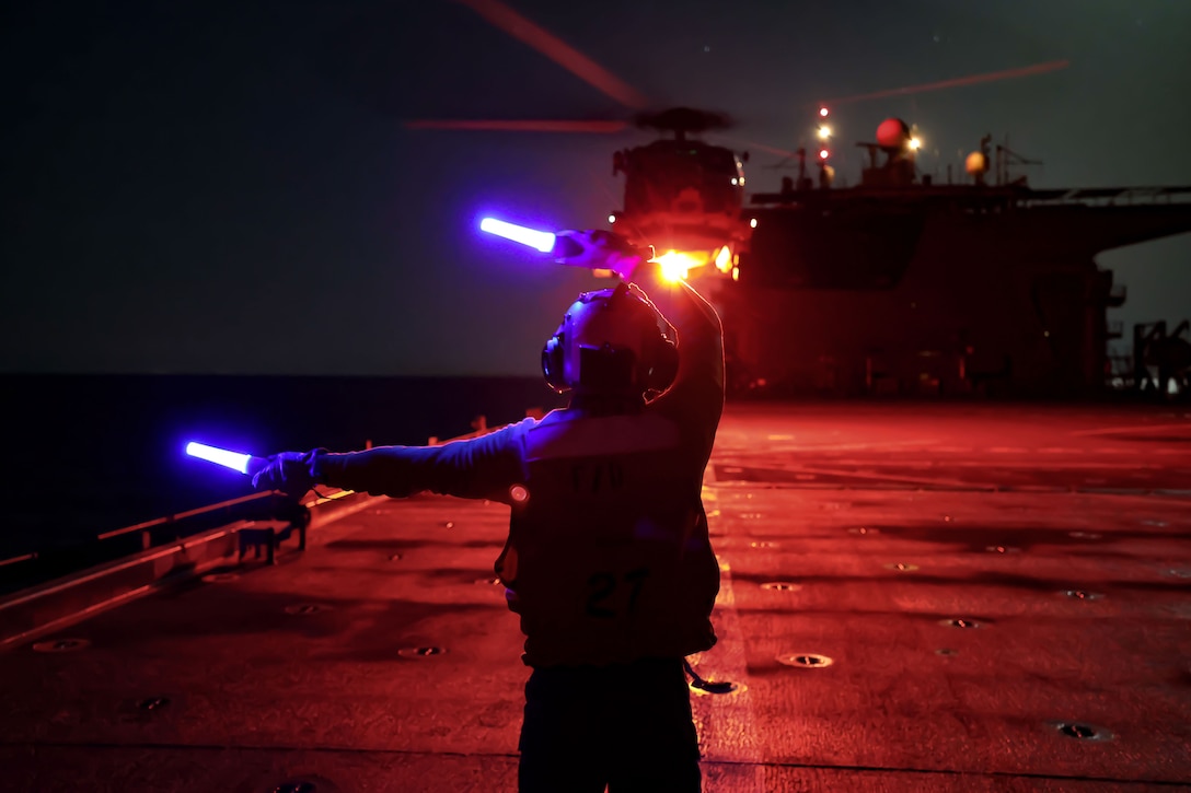 A sailor uses aircraft marshalling wands to direct a military helicopter on the flight deck of a ship at night.