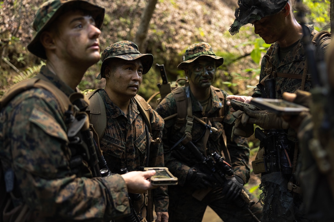 A group of Marines huddle for instructions in a wooded area while holding electronic devices.