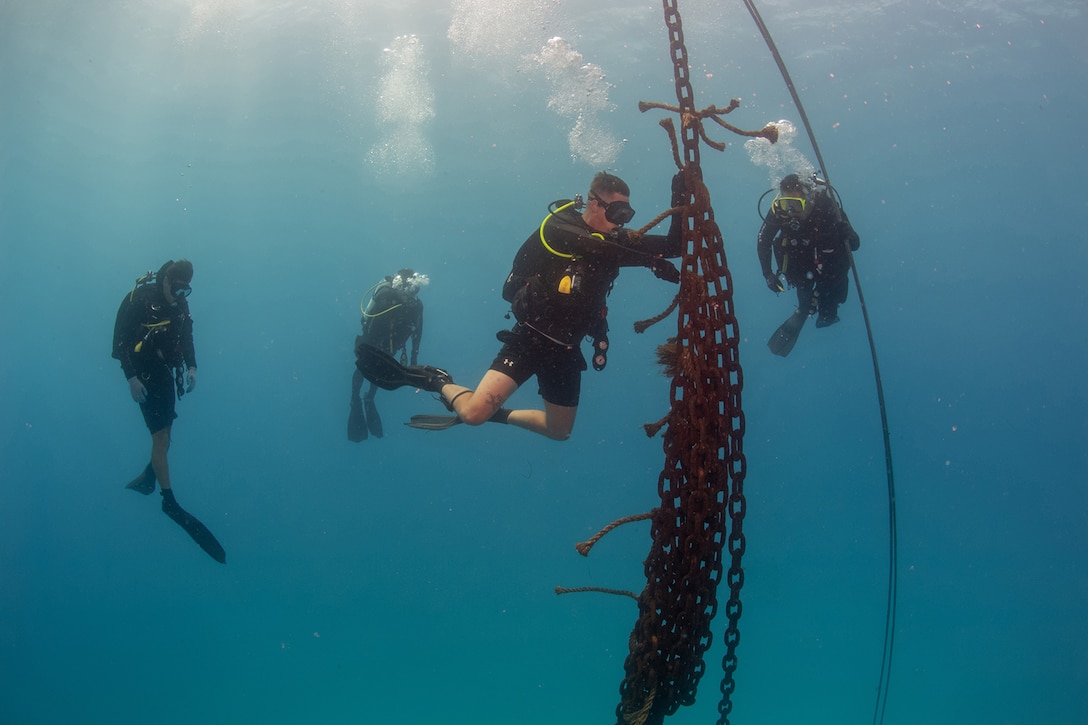 A Coast Guard diver cuts a line connected to chains underwater as other divers watch.