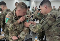 U.S. Army Spc. Trace Thompson, 11B, with the Missouri National Guard receives assistance from U.S. Army Sgt. Chadwick Larter, 79R, with the Massachusetts National Guard, in adjusting his competitor patch after receiving the welcome brief for the Army National Guard Best Warrior finals.