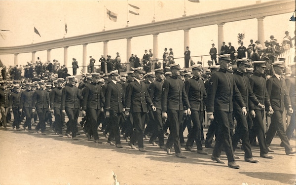 A group of men in uniform marching in column formation with an architectural feature visible in the background.