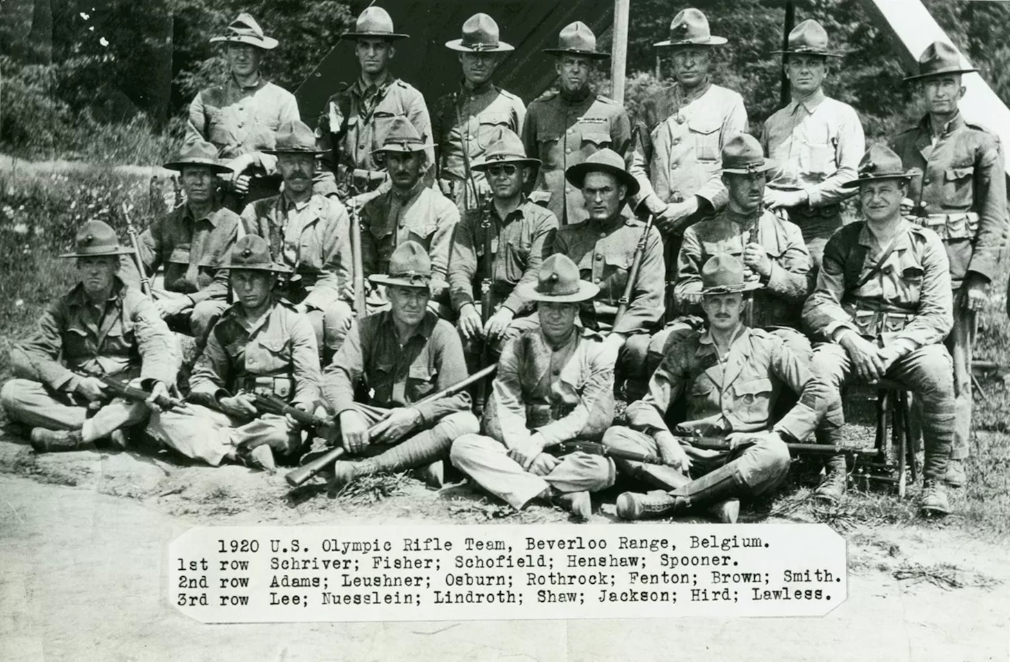 Three rows of men in khaki uniforms wearing brimmed hats. Some men are holding rifles.