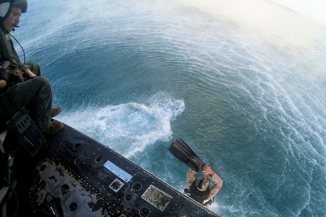 A Marine free falls into a body of water as a fellow Marine watches from a hovering helicopter.