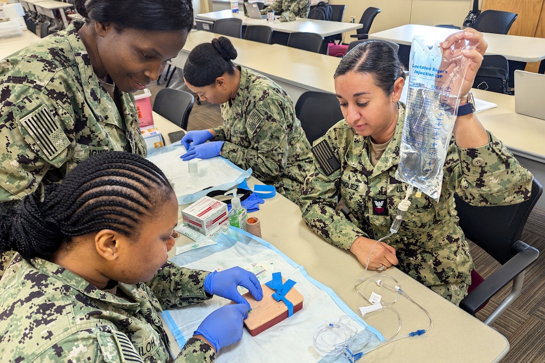 A sailor holds an IV bag as a fellow sailor cuts a square object on a medical pad while a fellow sailor watches.