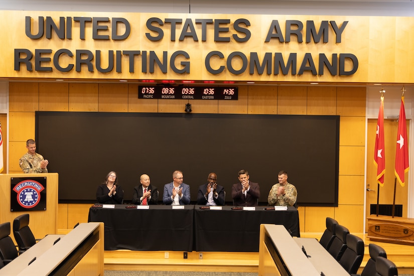 5 civilians sit across the front of the room with two U.S. Army Soldiers to sign a document for a training with industry partnership