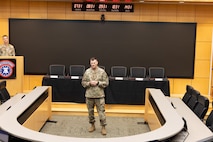 U.S. Army Soldier presenting behind a half circle desk with a long table and chairs behind him.