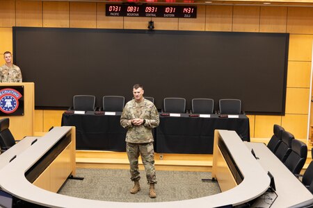 U.S. Army Soldier presenting behind a half circle desk with a long table and chairs behind him.