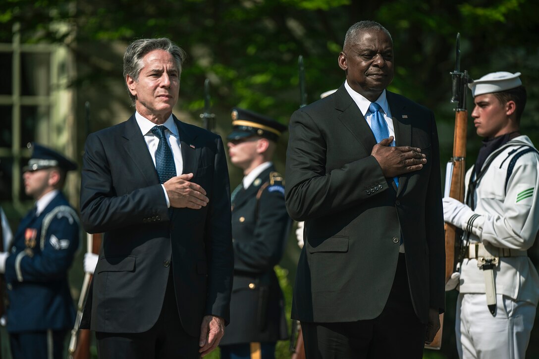 Two government officials in formal attire with right hands over heart stand at attention outside in a sunny day.