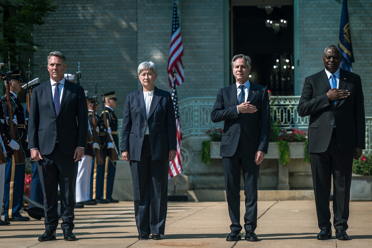 Four leaders stand outside, two with hands over hearts, with the American flag in the background and a military color guard off to the left.