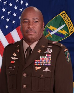 Chief Warrant Officer Five John Harris III poses in front of the American fliag and  U.S. Army Civil Affairs and Psychological Operations Command (Airborne). command flag in this command photo.