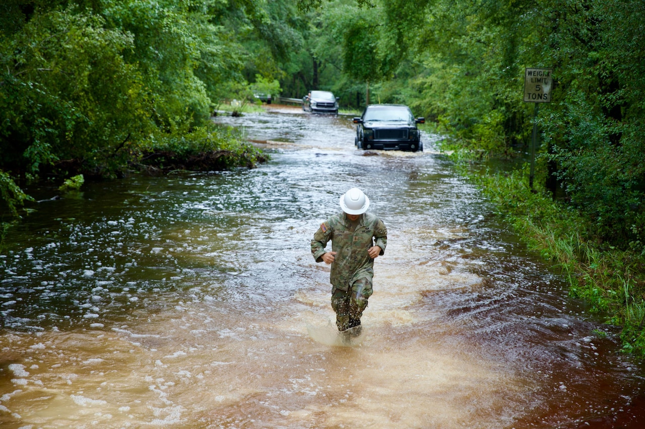 A soldier moves through flood waters with stranded vehicles behind and trees on either side.