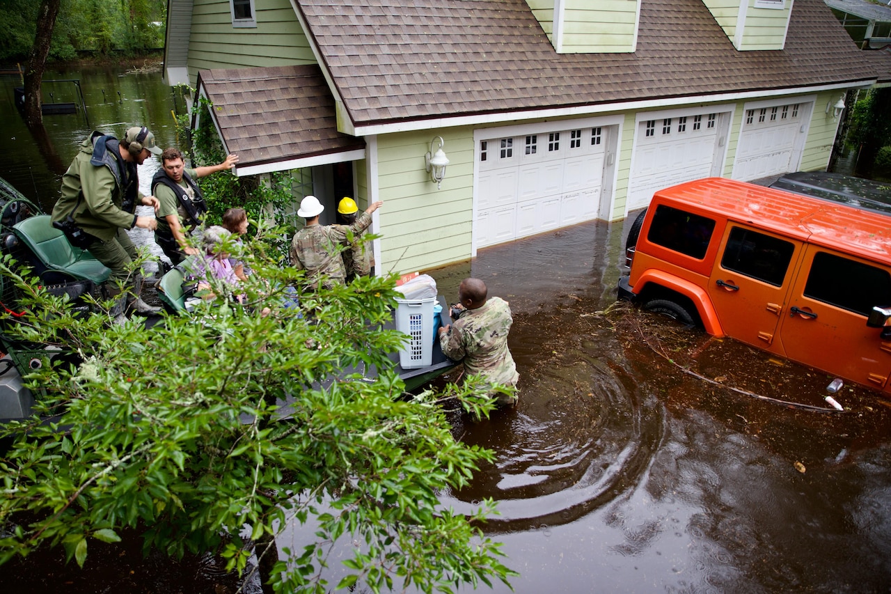 Five soldiers assist two residents evacuate a home with vehicles on the right.