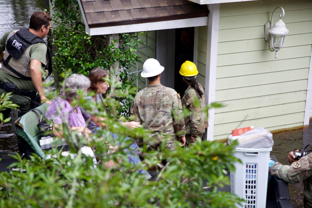 Three soldiers assist two residents by the porch of a house with shrubs on the foreground.