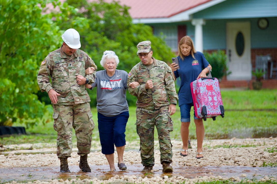 Two soldiers assist two residents through low flood waters with a home and shrubs in the background.