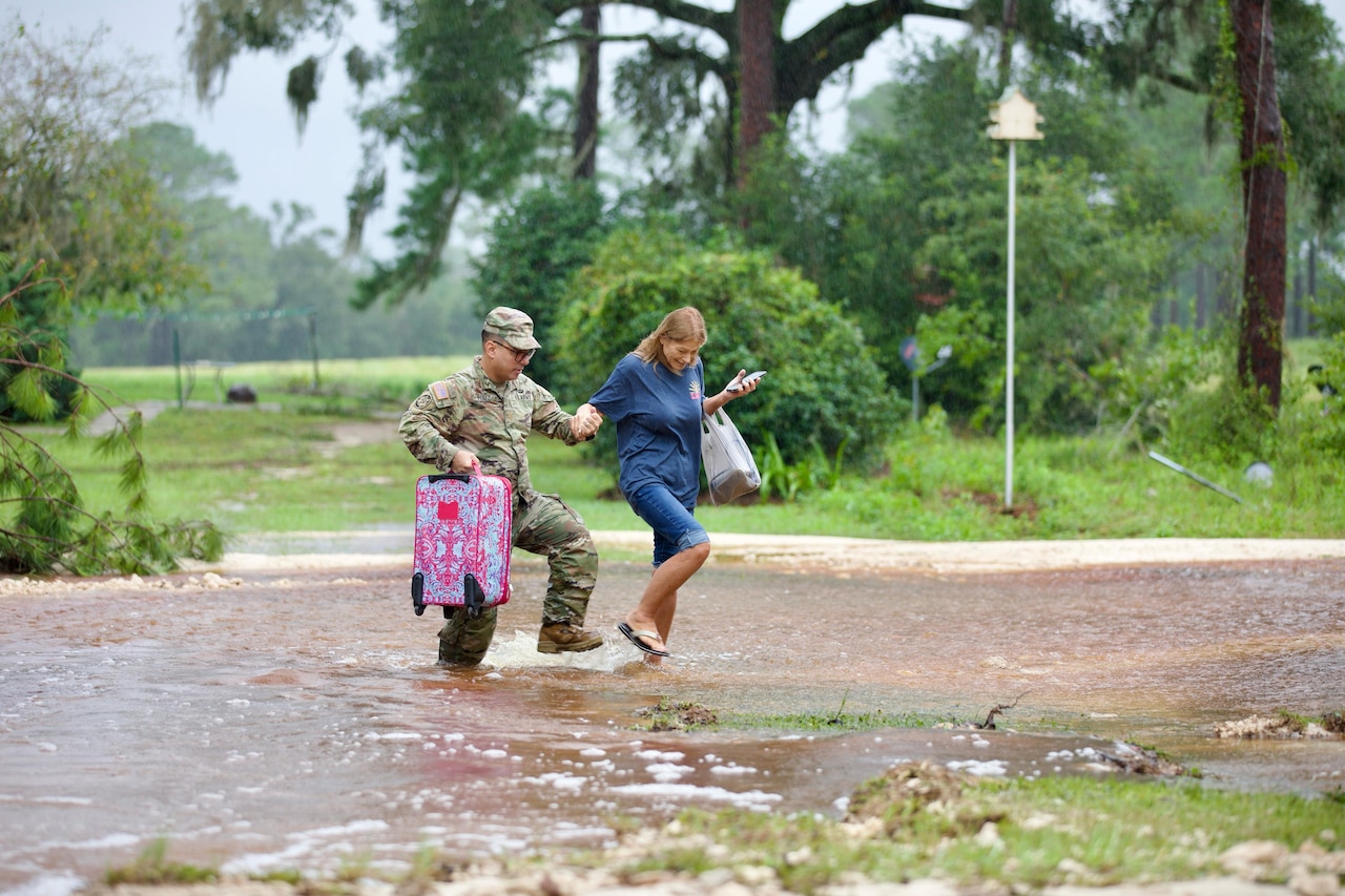 A service member assists a resident through low flood waters with trees in the background.