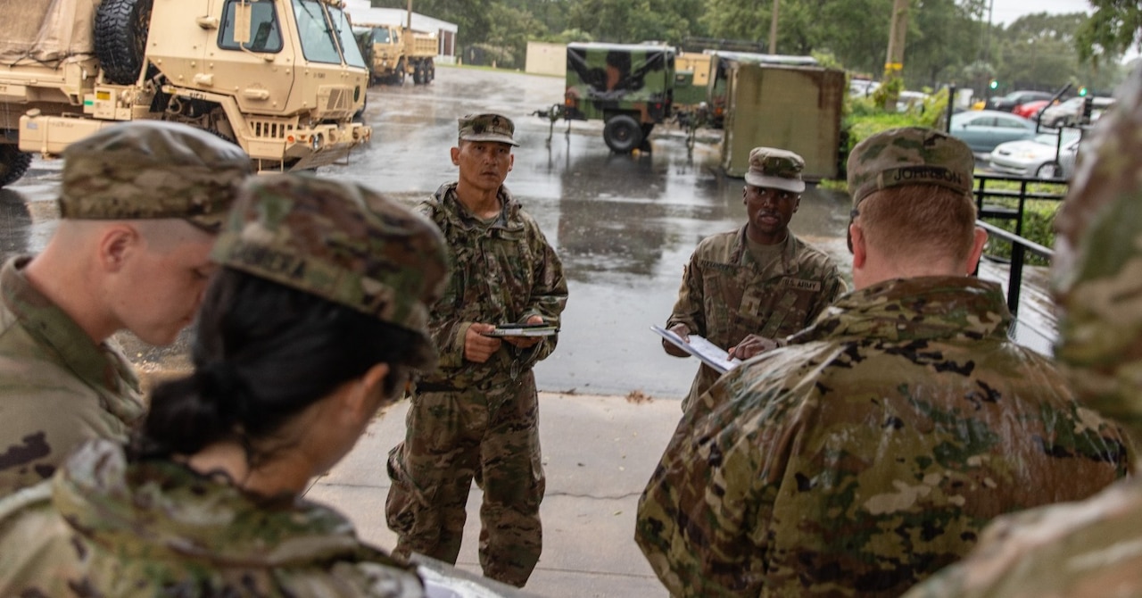 A group of guardsmen stand outside in discussion with military vehicles in the background.