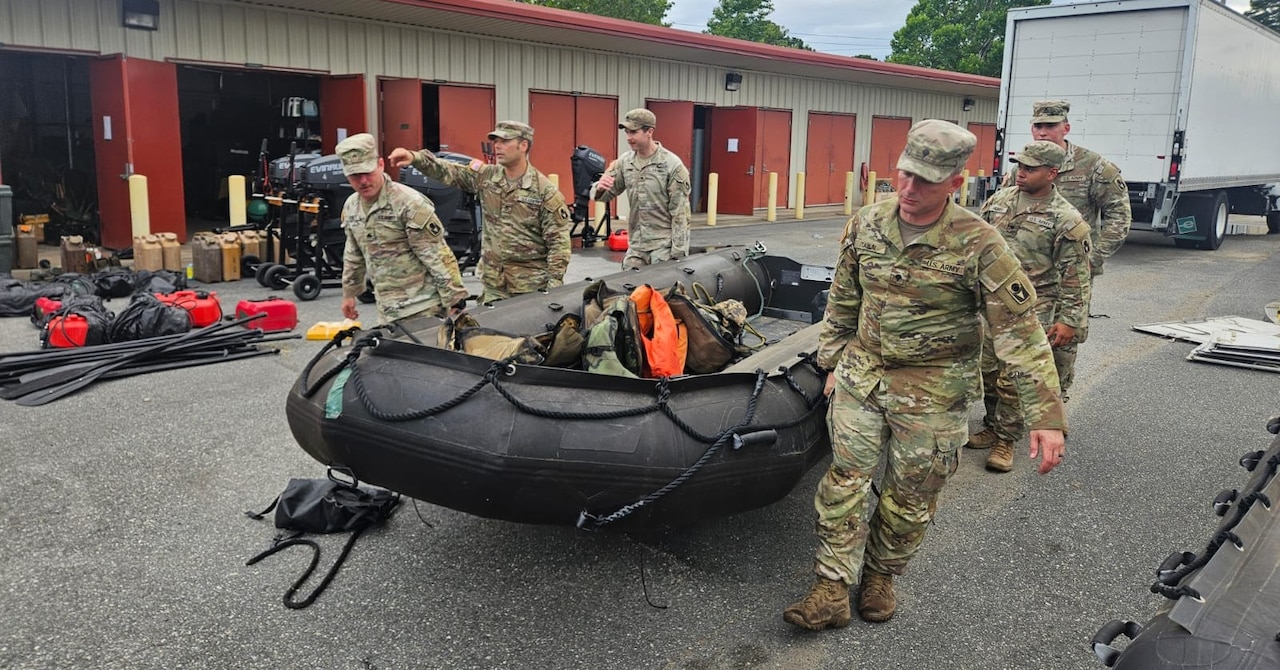 Several soldiers carry an inflatable rubber craft alongside a storage building with a vehicle in the background.