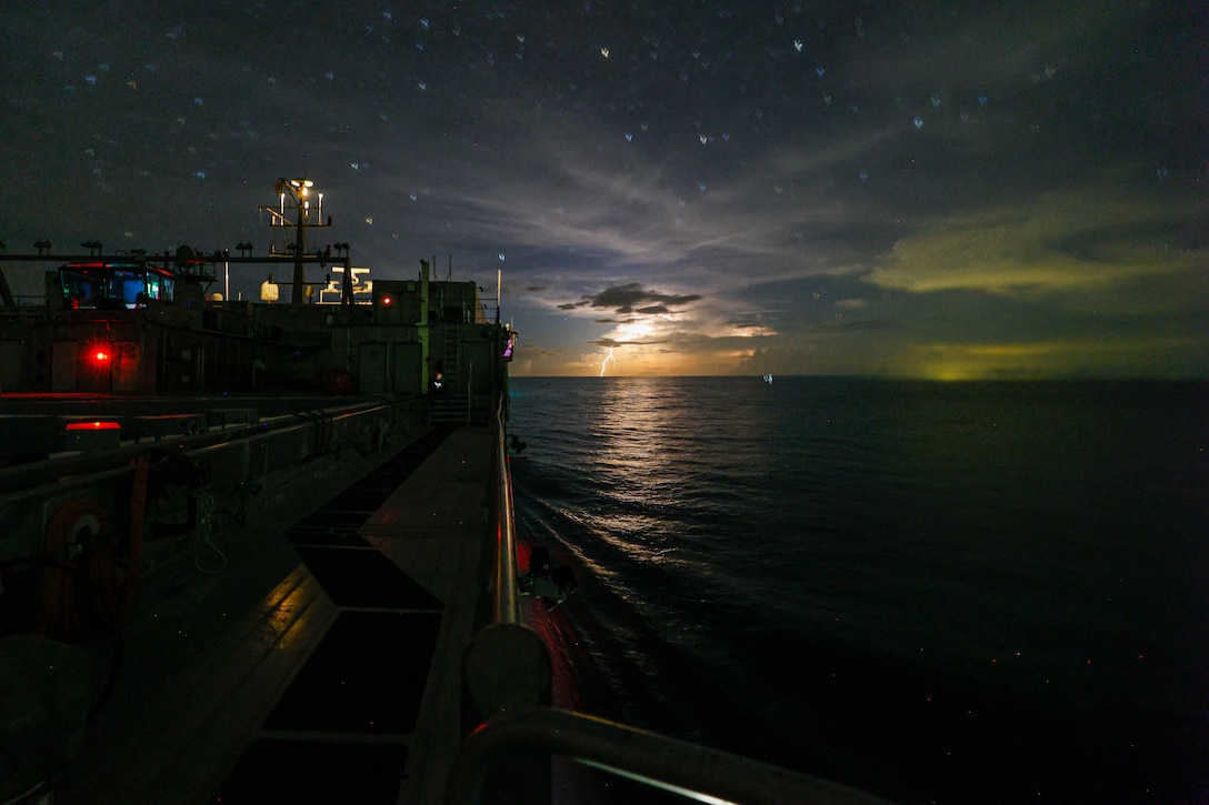 A ship steams through the water at night under a starry sky with lightning flashing on the horizon.