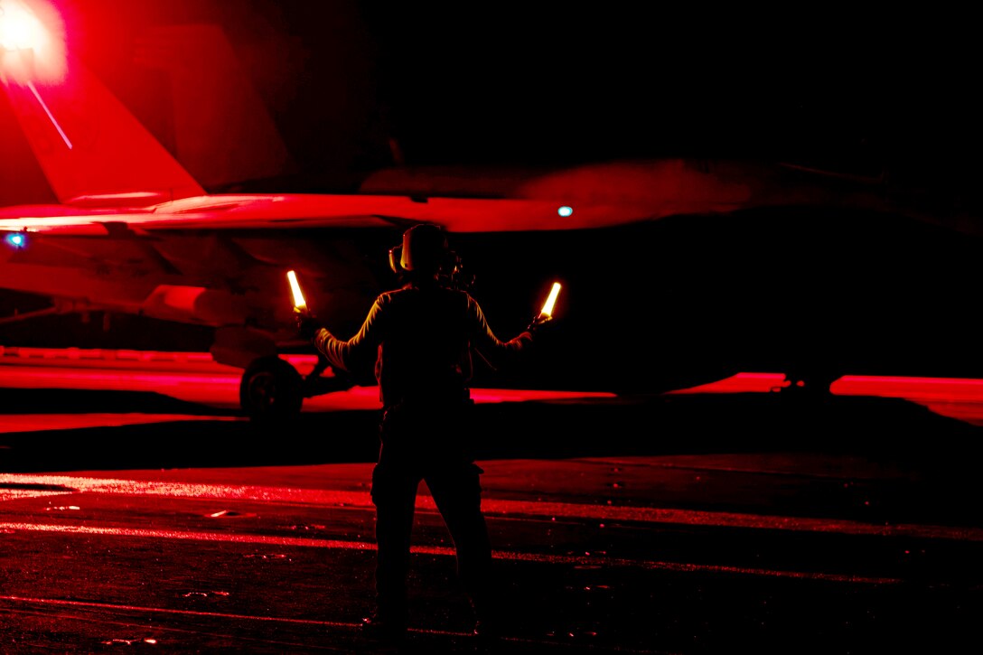 A sailor uses marshalling wands to guide an aircraft aboard a ship under red lights at night.