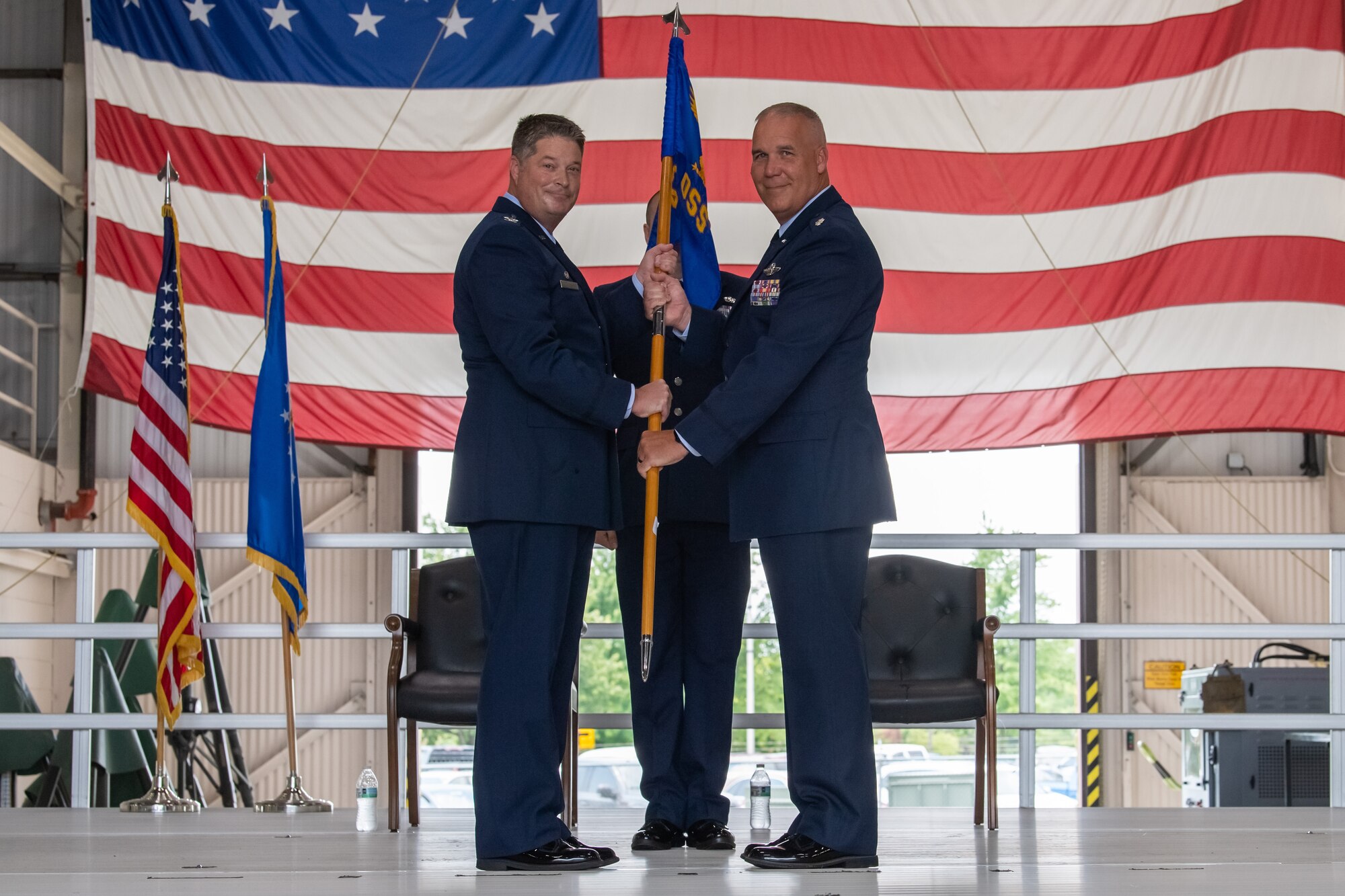 Two men in U.S. Air Force dress uniforms pose for a photo while holding a guidon. In the background, an American flag.