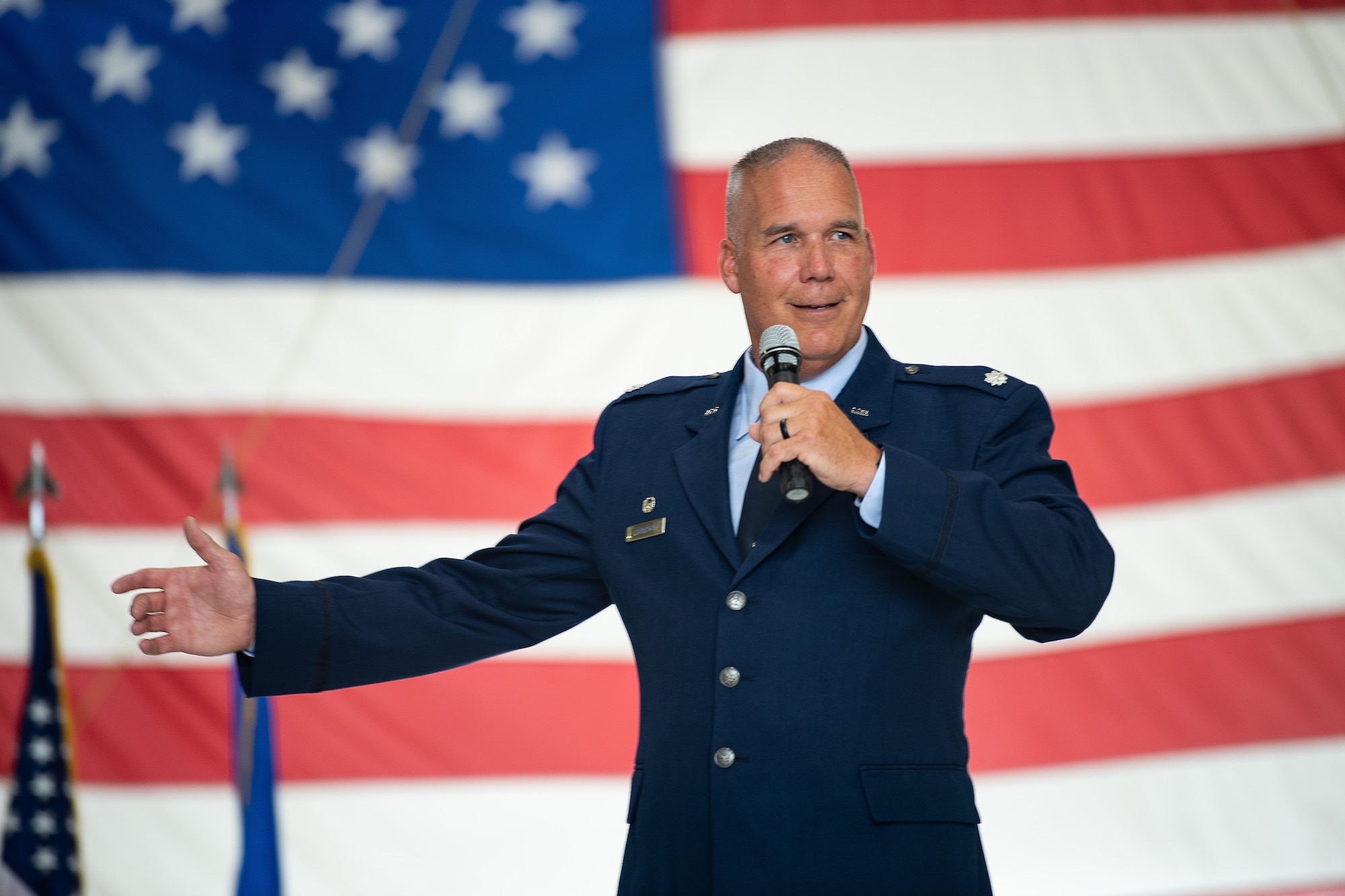 A man in a U.S. Air Force dress uniform talks into a microphone he is holding while gesturing with his other arm.