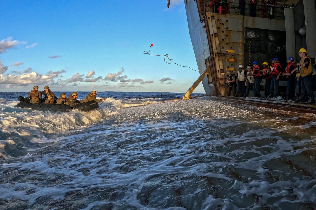 Service members in a raft approach a group of service members standing inside the loading dock of a Navy ship during daylight.