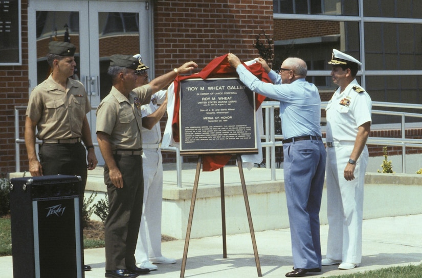 A person in civilian clothing lifts a sheet of a plaque with two service members as two others in uniform stand by.