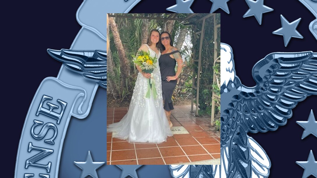 Woman in a whit wedding dress standing while holding yellow flowers in her right hand with her left are wrapped around her mother in a dark dress.