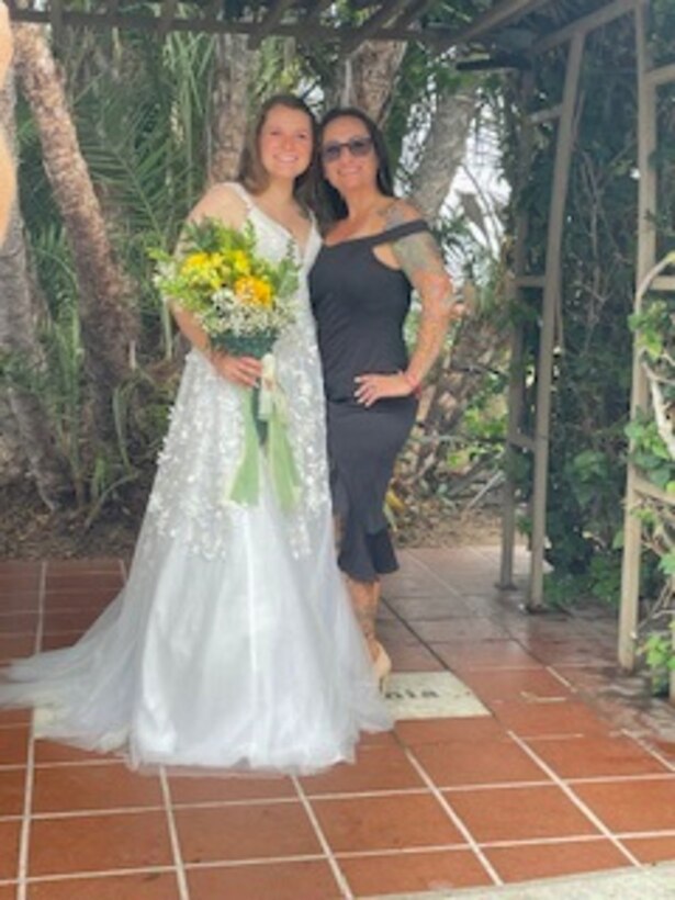 Woman in a whit wedding dress standing while holding yellow flowers in her right hand with her left are wrapped around her mother in a dark dress.