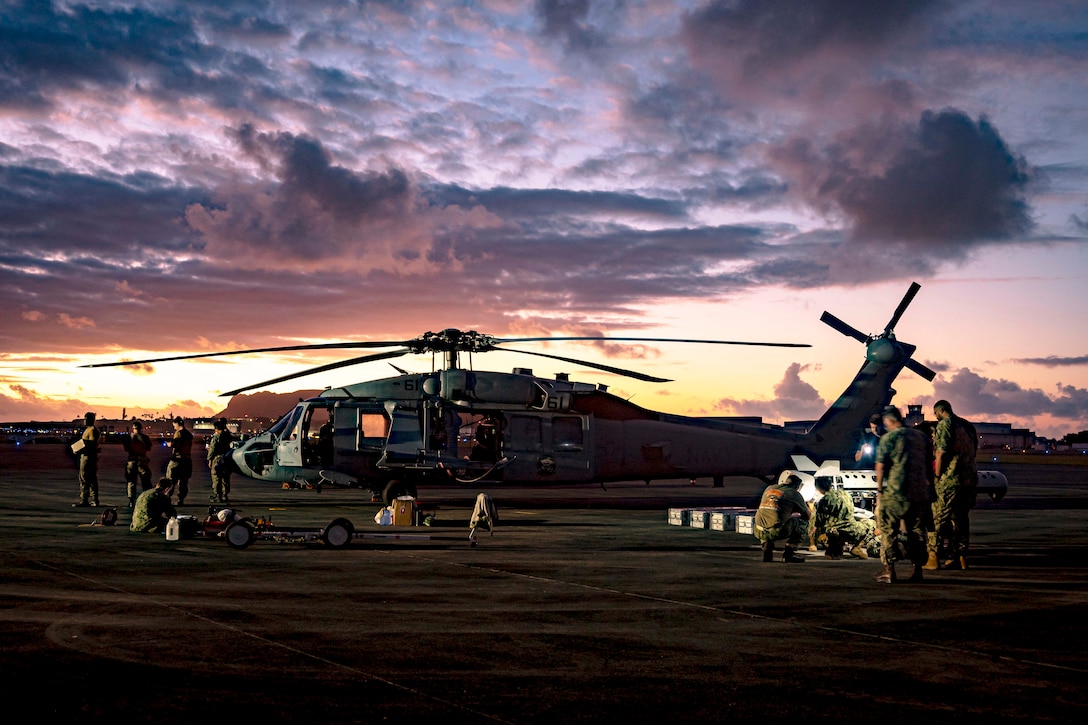 Small groups of sailors gather on a tarmac around a parked helicopter under a low sitting sun.